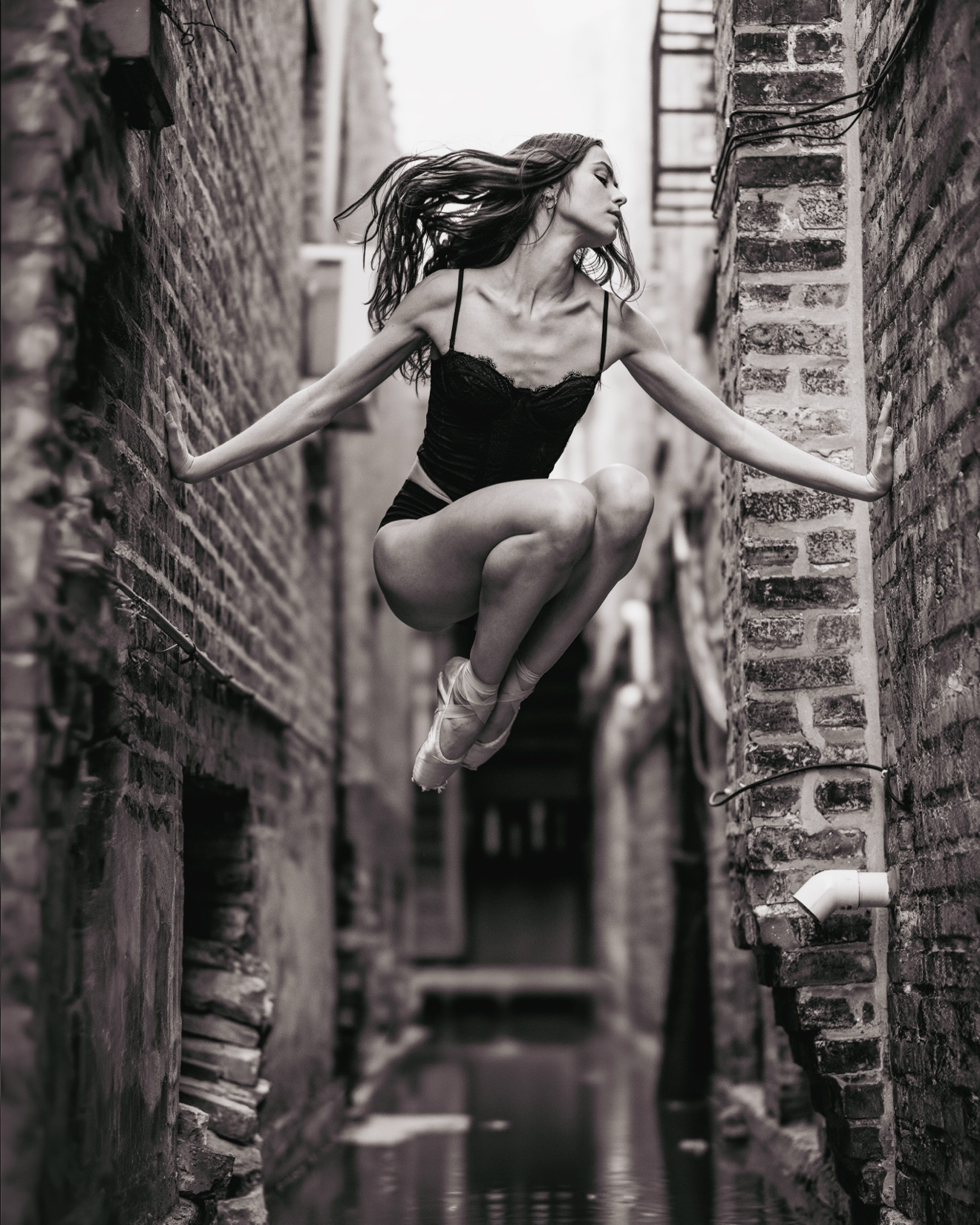 A ballet dancer suspended mid-jump between two brick walls in a Chicago alley, hands touching both walls, black and white